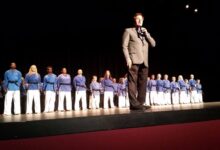Stephen Oliver speaking on stage in front of a line of Mile High Karate black belt students