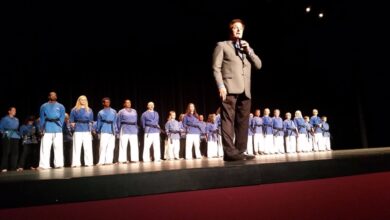 Stephen Oliver speaking on stage in front of a line of Mile High Karate black belt students