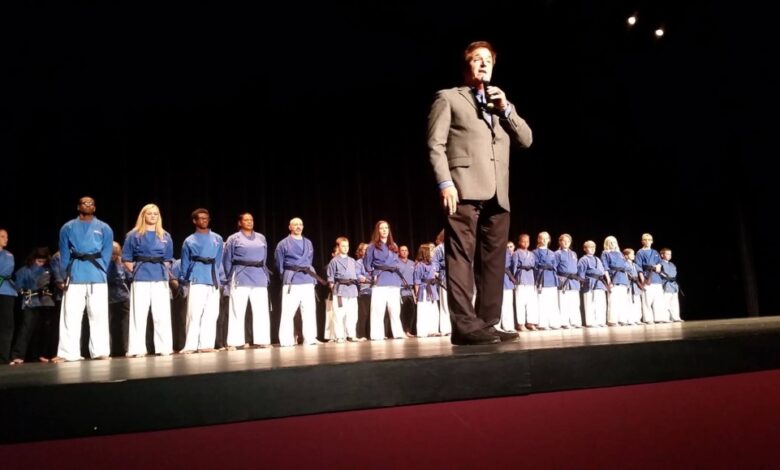 Stephen Oliver speaking on stage in front of a line of Mile High Karate black belt students