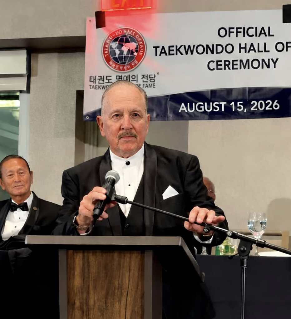 A speaker in black tie at the podium beneath the Official Taekwondo Hall of Fame Ceremony banner dated August 15, 2026