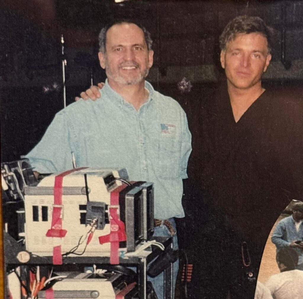 Stephen Oliver in a black uniform with Ed Saenz beside camera equipment on the set of Walker, Texas Ranger